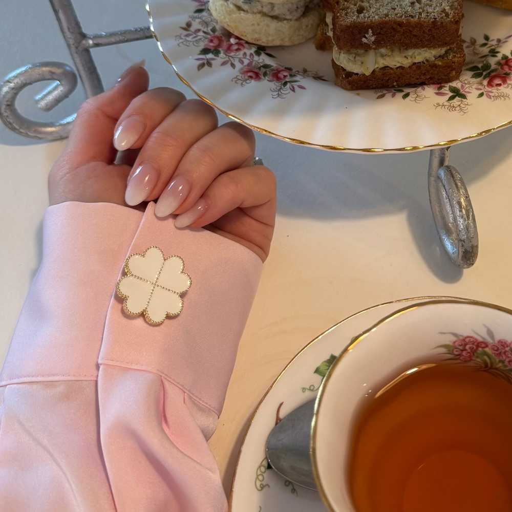 A close-up lifestyle shot of a woman's hand wearing a light pink satin sleeve with a decorative white clover-shaped decorative button cover on a sleeve (cuff. Romantic, elegant and modern, set next to a cup of tea and a floral cake stand with sandwiches.