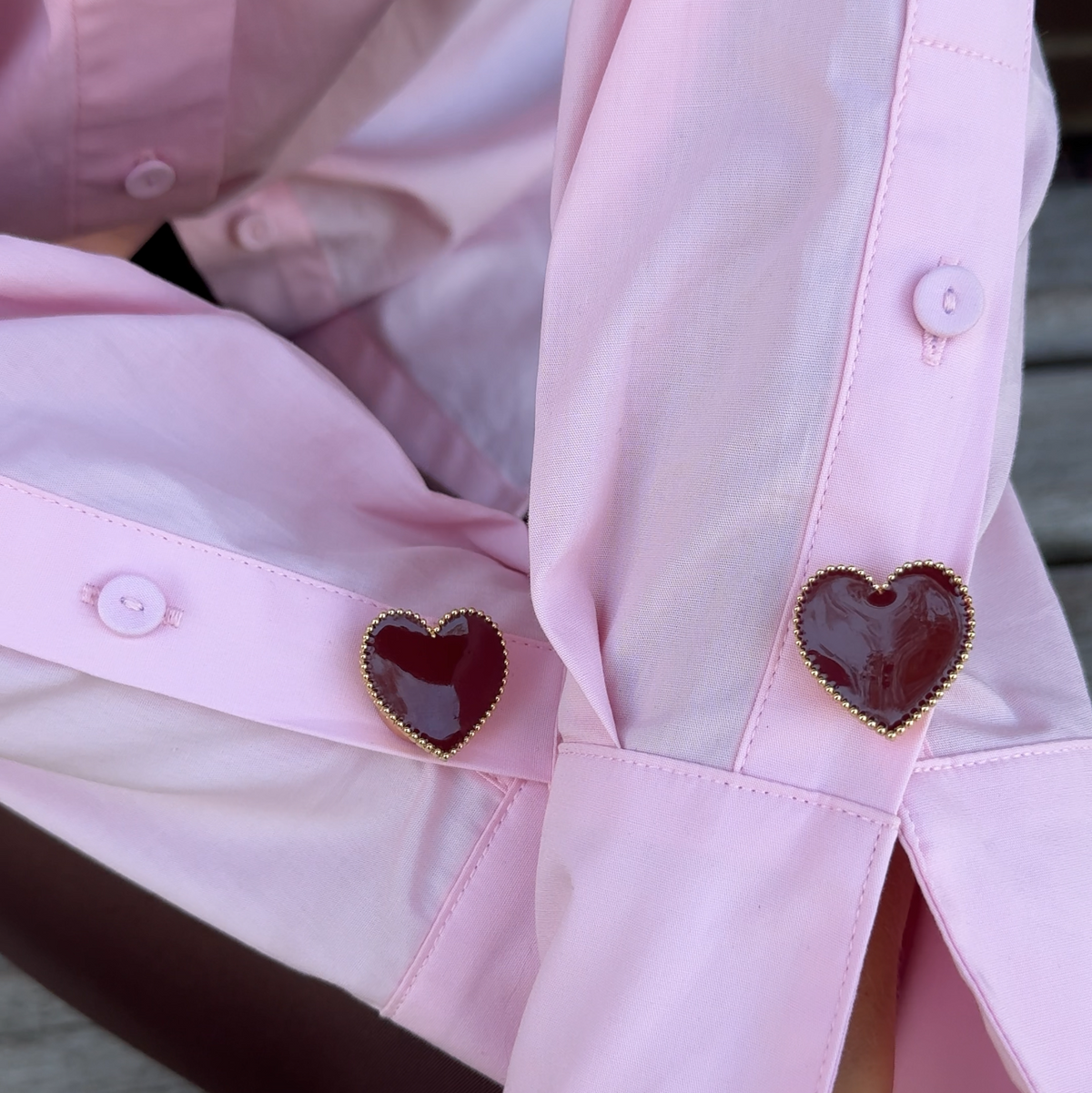 A close-up of a light pink dress shirt with long sleeves with decorative heart-shaped gold-dotted button covers. The buttons have dark merlot and burgundy like resin centers
