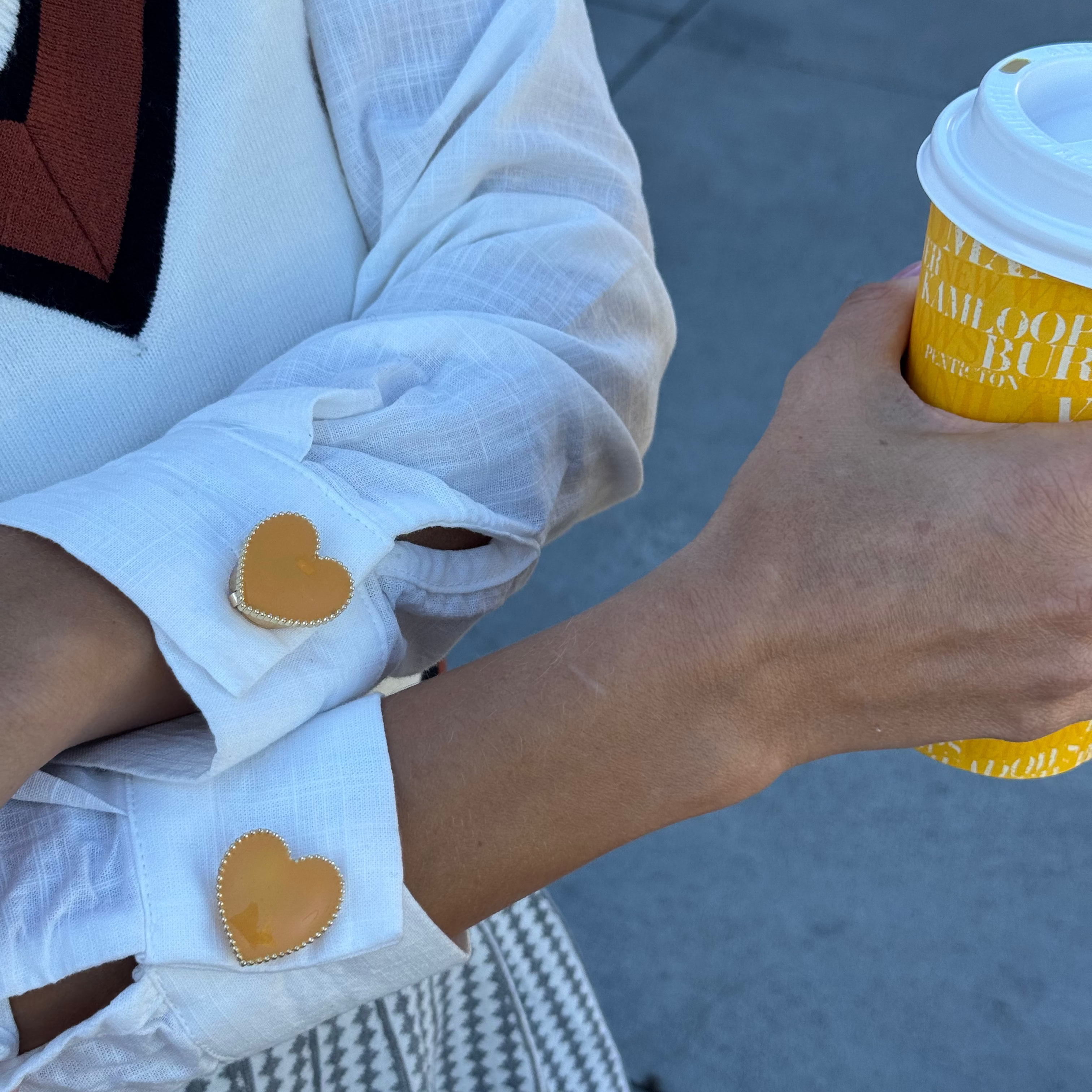 A close-up lifestyle shot of a person wearing a white linen long sleeve shirt with mustard color heart-shaped gold-dotted decorative button cover on each sleeve (cuff), holding a yellow coffee cup outdoors on a gray paved surface.