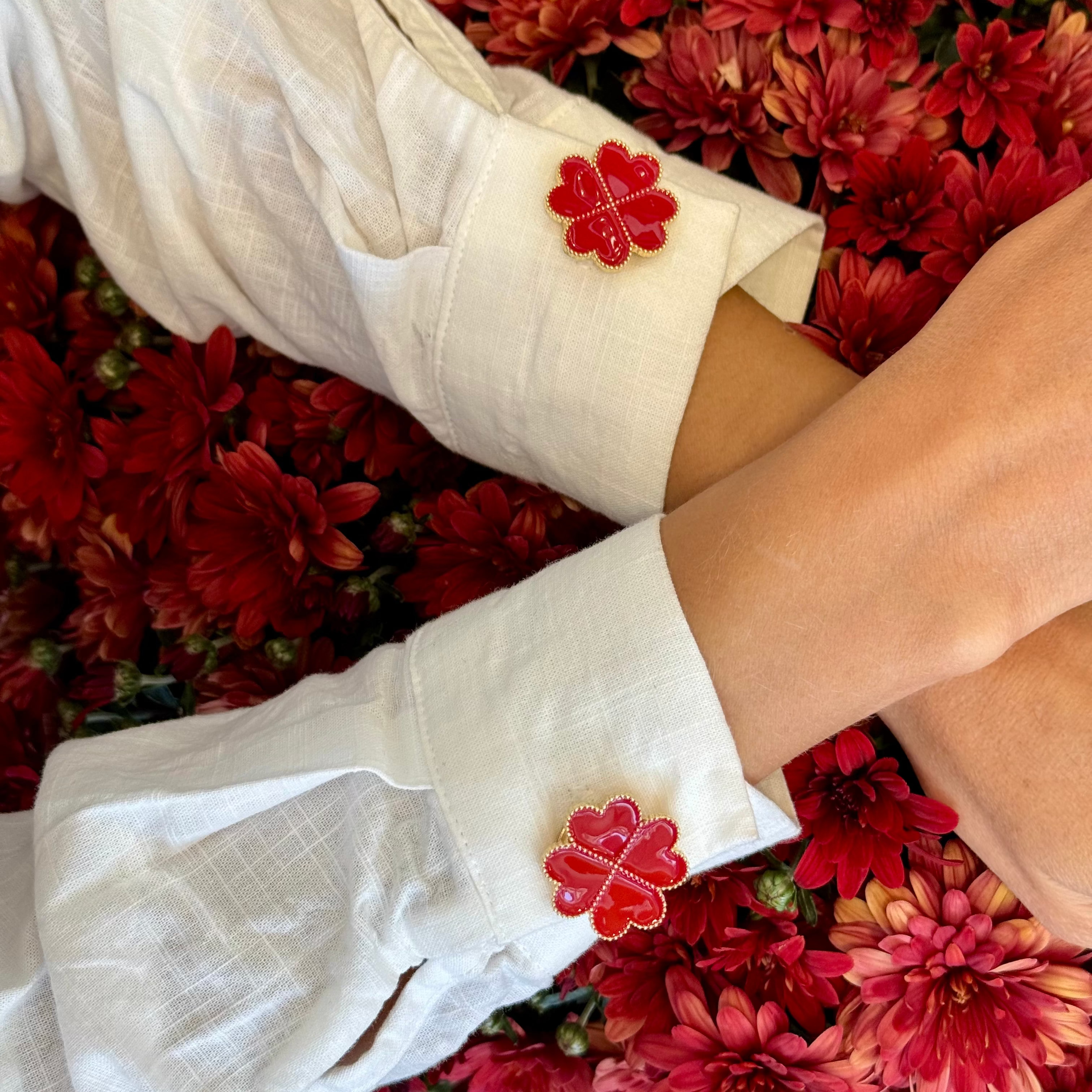 A close-up of a person wearing a white linen shirt with two red four-leaf clover decorative button cover on a sleeve (cuff). Romantic, elegant and modern, set against a dense bed of red flowers.
